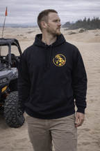 Man wearing a black hoodie with Naughty Goat logo in a desert-like setting with off-road vehicle.