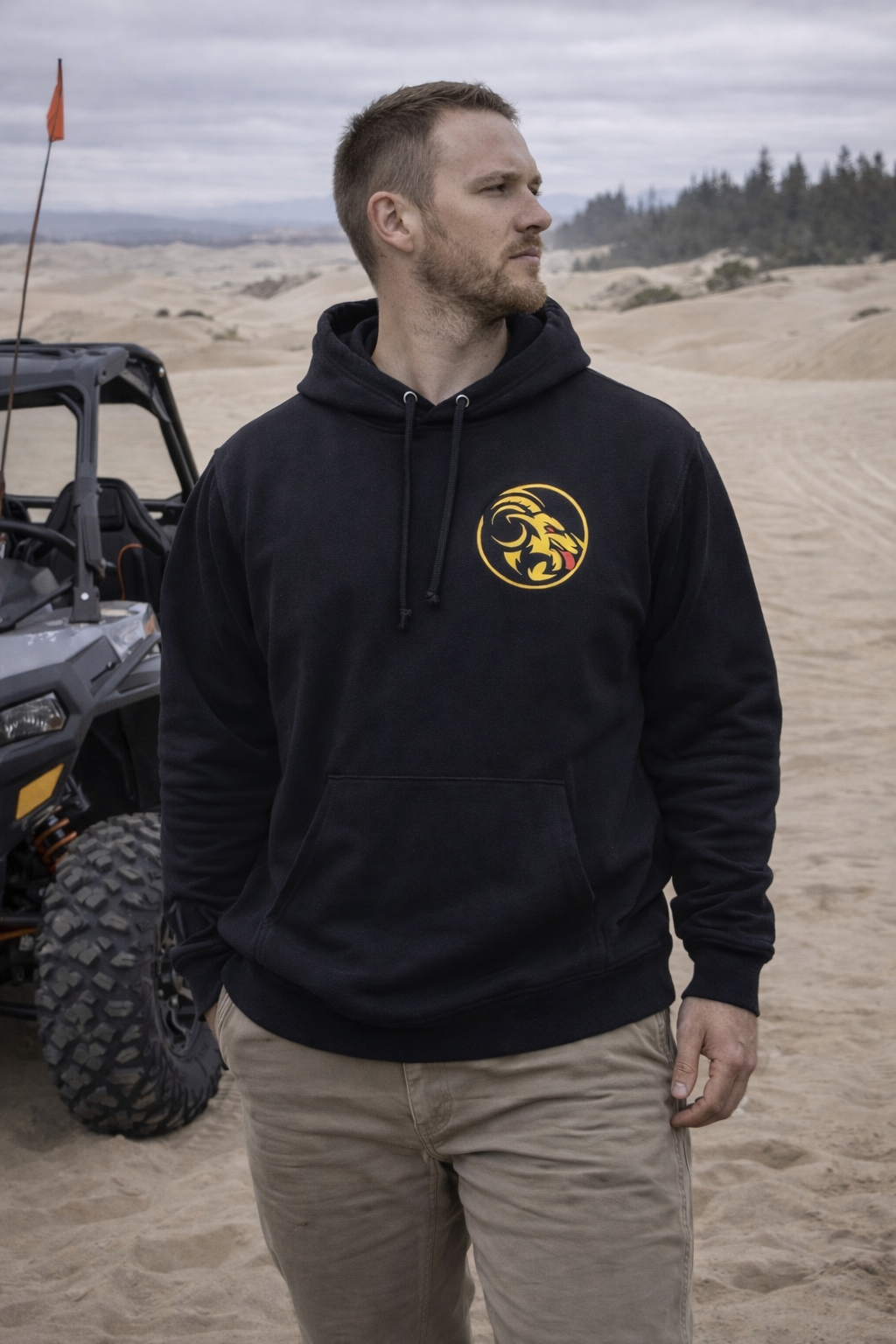 Man wearing a black hoodie with Naughty Goat logo in a desert-like setting with off-road vehicle.