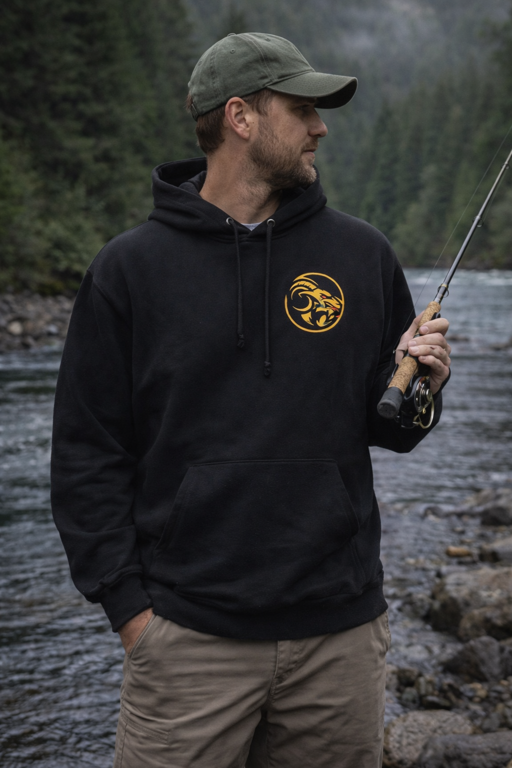 Man wearing a black hoodie with Naughty Goat logo, holding a fishing rod by a river.