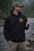 Man wearing a black hoodie with Naughty Goat logo, holding a fishing rod by a river.