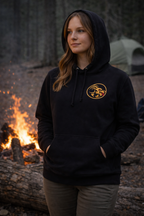 Person wearing a black hoodie with Naughty Goat logo in front of a campfire and tent in a forest setting
