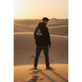 Person wearing a black hoodie with a logo standing in a desert at sunset.